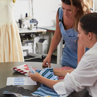 Two designers in a fashion workshop setting, one pointing at fabric swatches on a table.